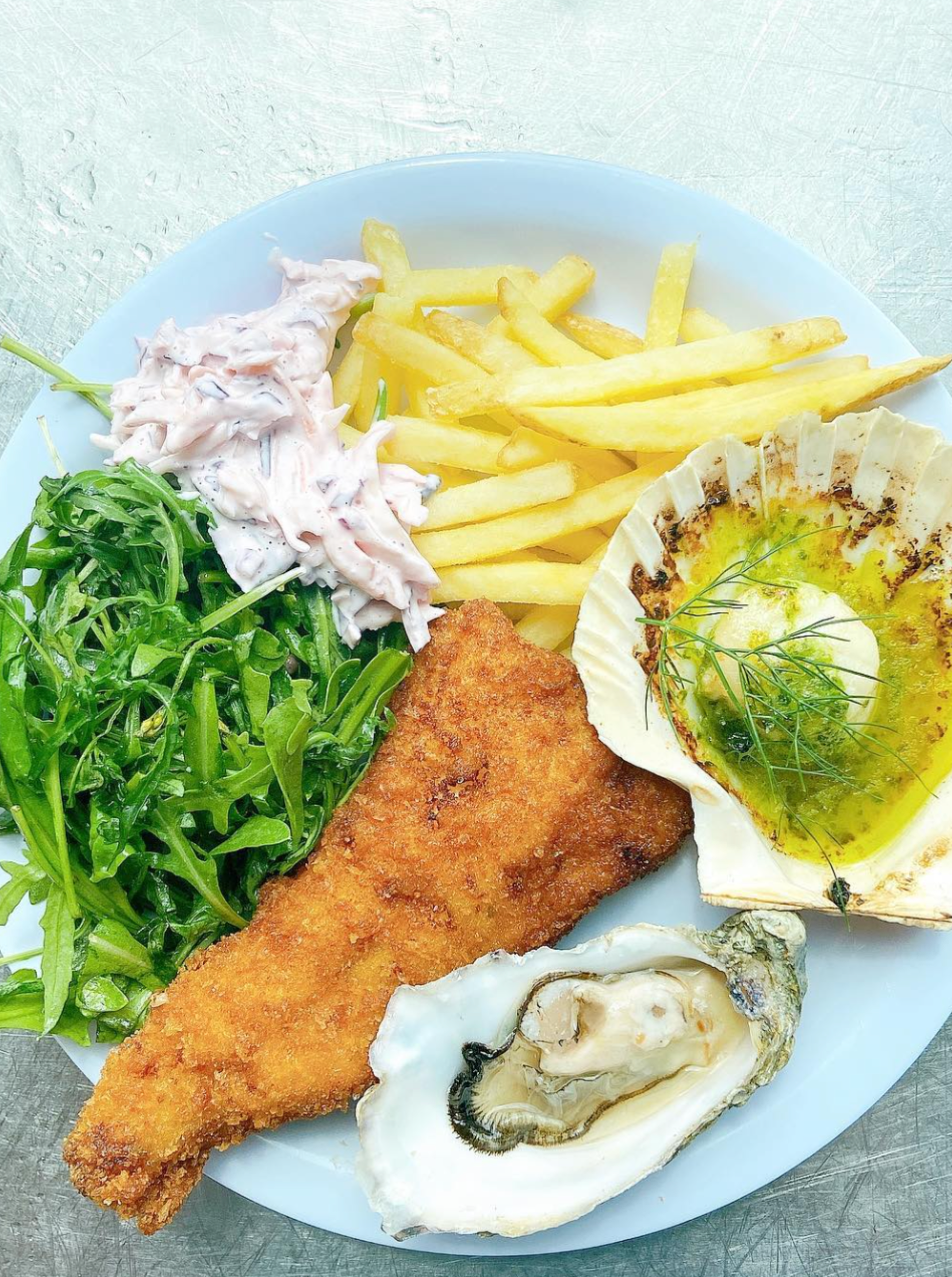 A plate of food is shown: Breaded fish fillet, chips, coleslaw, salad, an oyster and a mussel.