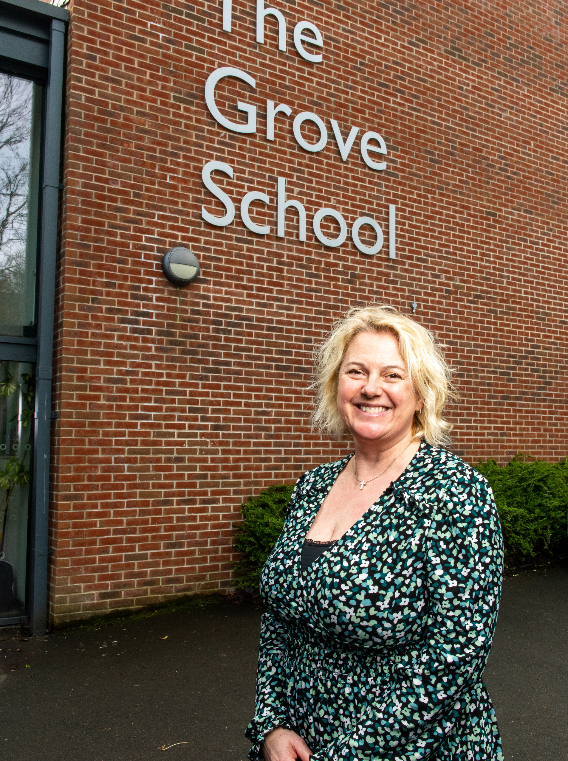 Headteacher Hilary stands infront of the school sign that says "The Grove School" smiling